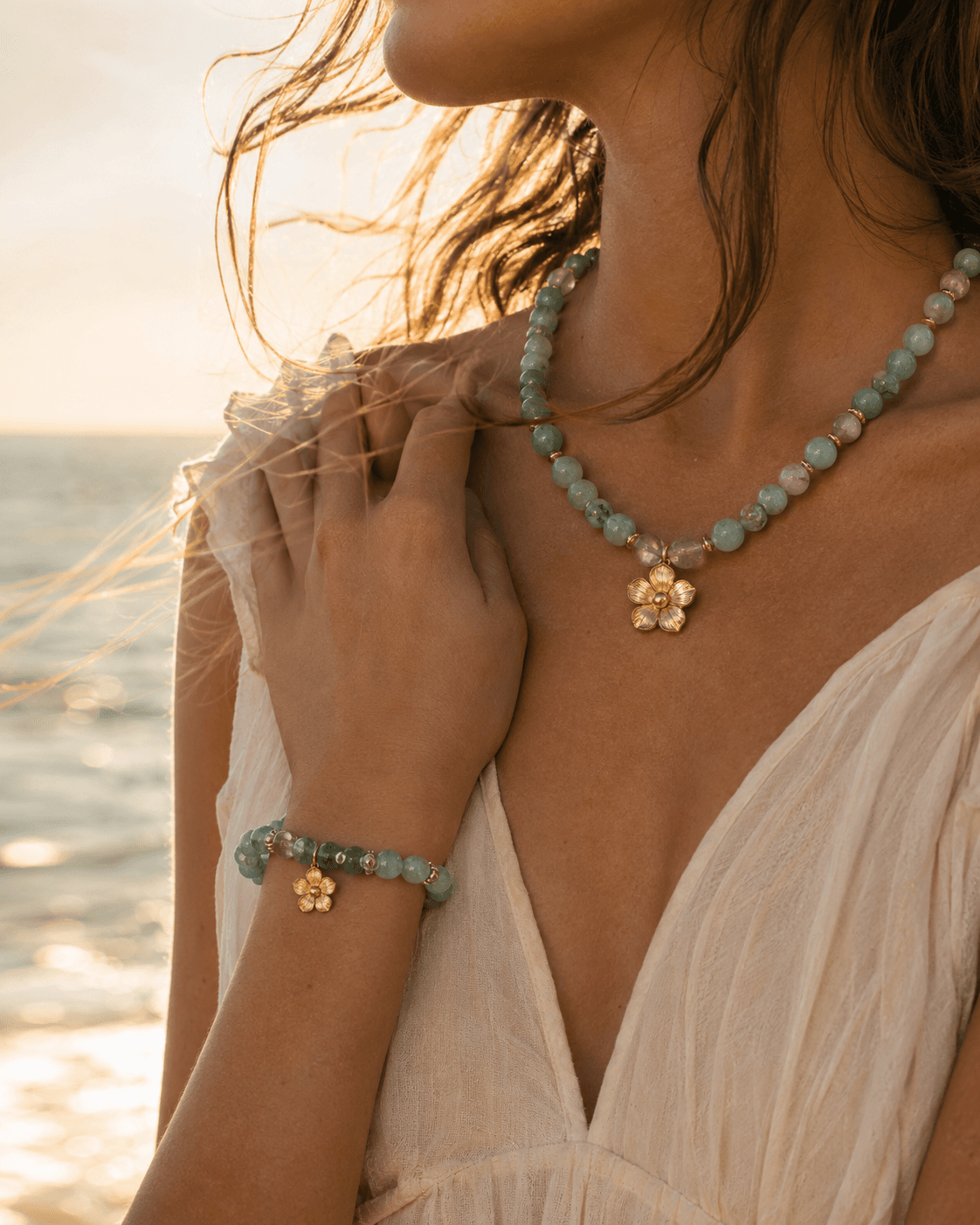 Mujer en vestido de lino color marfil junto al mar luciendo un collar y una pulsera de aguamarina con dijes de flor dorada
