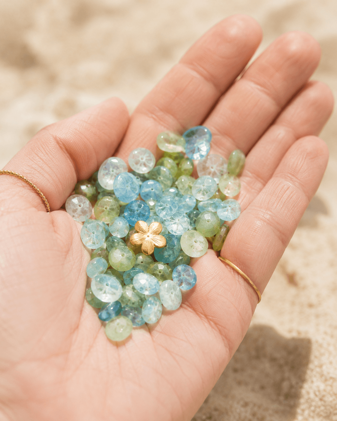 Mano sosteniendo un puñado de cuentas de piedras naturales en tonos aguamarina, jade y celeste sobre la arena