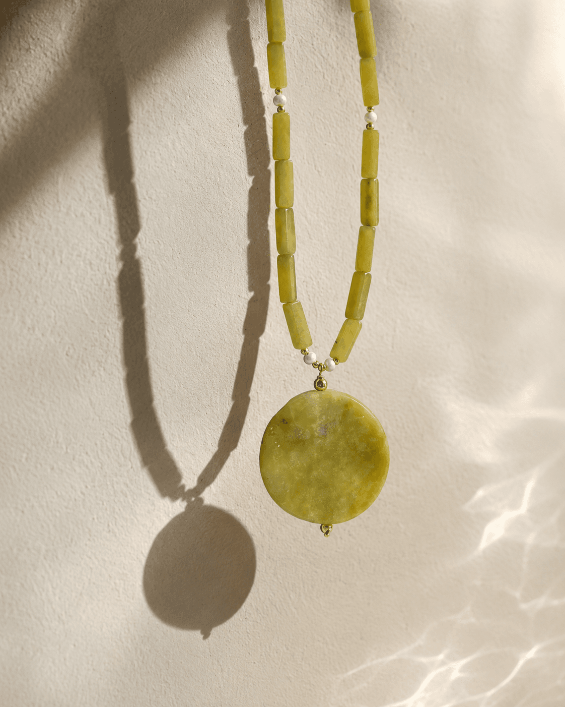 Collar de jade limón con medallón circular colgando frente a una pared clara con sombra.