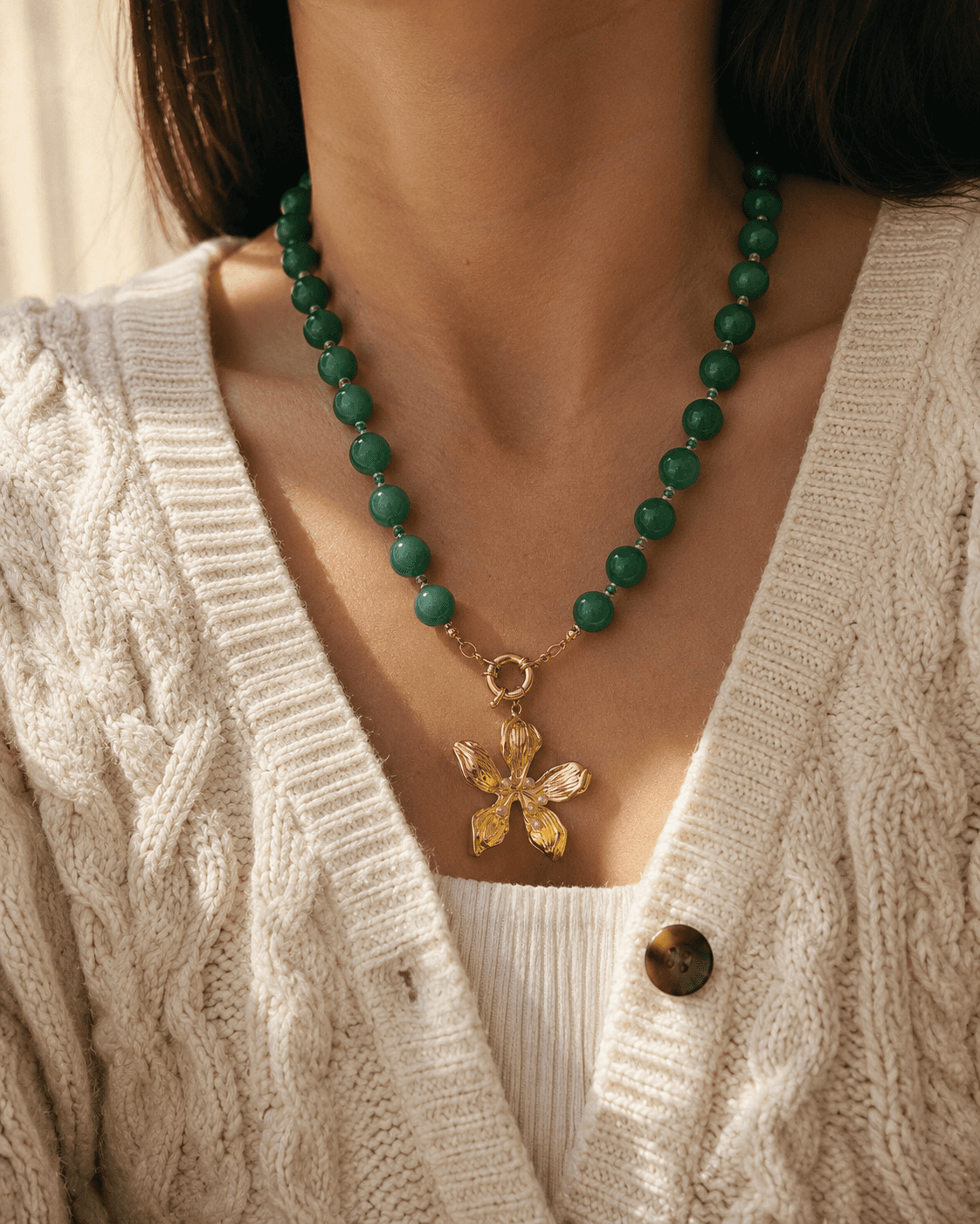 Collar de cuentas de aventurina verde con dije de flor dorada sobre cuello de mujer con chaleco de lana crema.