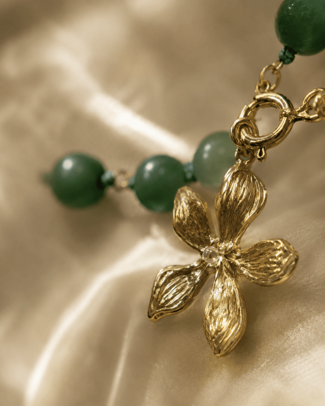 Detalle macro del dije de flor de cinco pétalos en baño de oro junto a cuentas de aventurina verde.