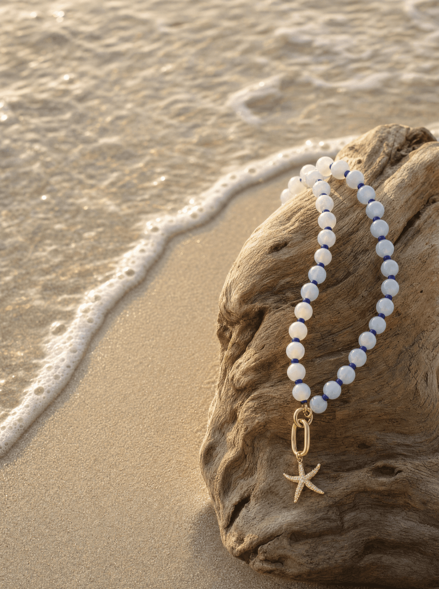 Pulsera con estrella de mar dorada sobre madera de deriva a la orilla del mar al atardecer.